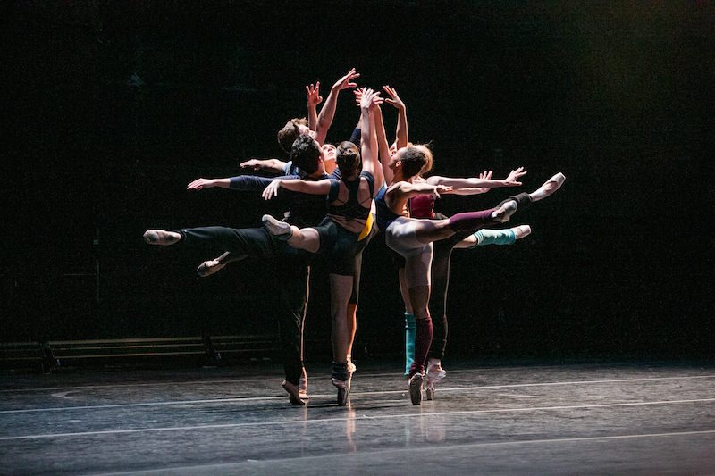 Dancers in colorful leotards and leg warmers pique in arabesque facing into a small circle